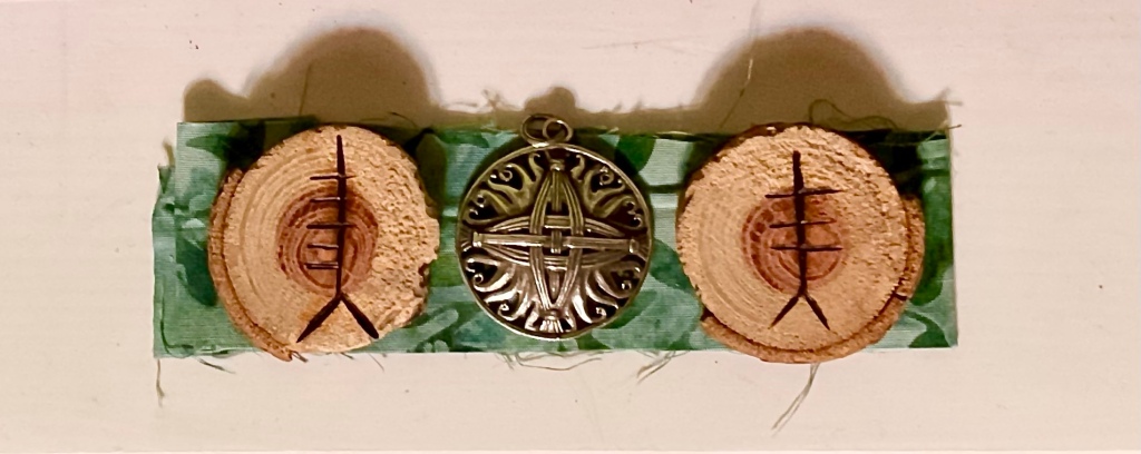 On a strip of variegated green fabric, three round objects: a wooden token inscribed with the ogham symbol Ceirt (five short horizontal lines extending leftward for a vertical stroke), a silver medallion in the shape of an interwoven Brigid’s cross, and another wooden token with the ogham symbol Úr (three short horizontal lines crossing perpendicularly across a vertical stroke)