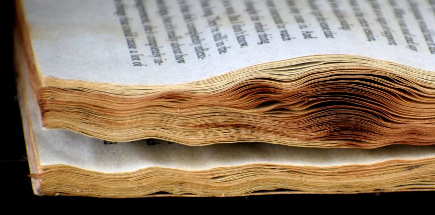 Side view of a water-damaged book with text blurred. The page edges are dark yellow and irregularly wavy.